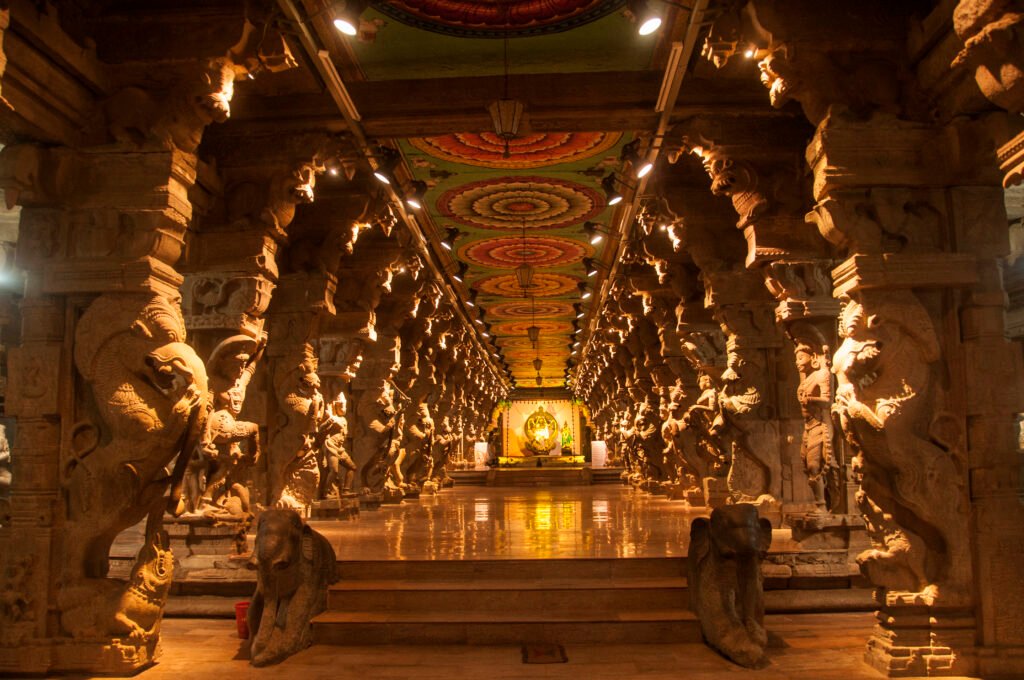 Ornate interior corridor of a South Indian temple featuring intricately carved stone pillars, colourful ceiling artwork, and a glowing shrine at the far end.