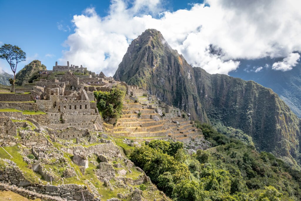 Panoramic view of Machu Picchu’s ancient Inca stone terraces and ruins set against steep green mountains and a bright cloudy sky.